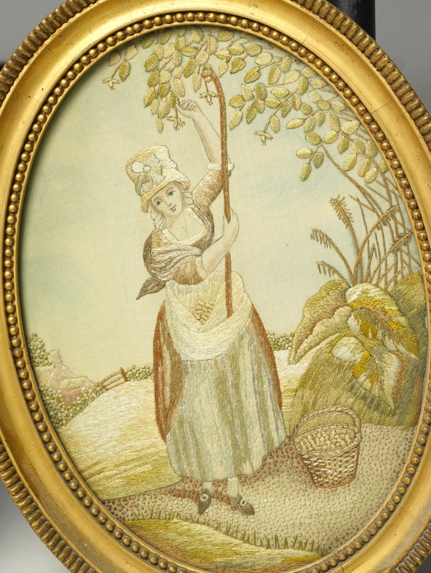 A pair of early 20th century small gilt oval embroideries, of a girl gathering fruit from a tree and a seated lady in a landscape, worked in stem stitch and decorative knotting. 29cm long including gilt frames. Condition